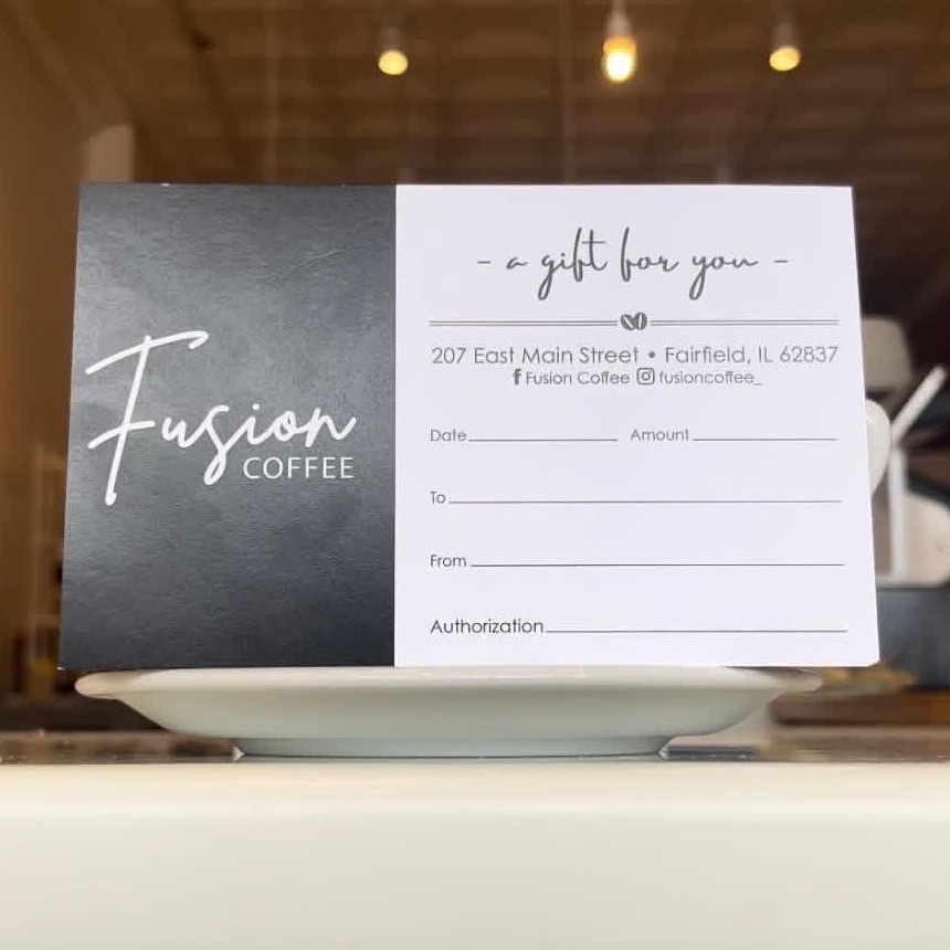 Fusion Coffee gift card — charcoal logo panel beside a “a gift for you” card — propped on a saucer on the counter.